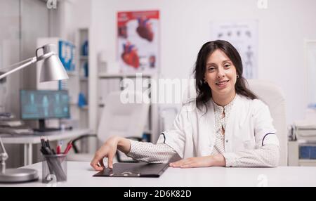 Fröhliche attraktive Frau Arzt im Krankenhaus Büro trägt weißen Mantel lächelnd vor der Kamera. Stockfoto