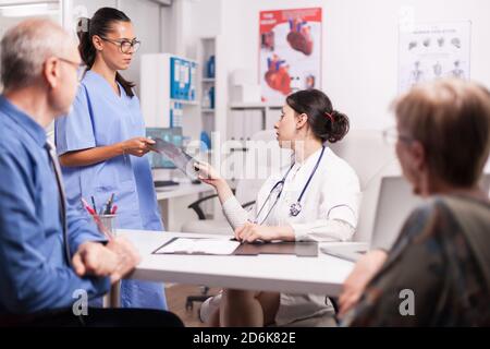 Krankenschwester mit Brille, die Arzt Röntgen während der Konsultation der kranken verheirateten Rentner im Krankenhaus Büro. Junge Sanitäter mit weißem Mantel und Stethoskop. Stockfoto