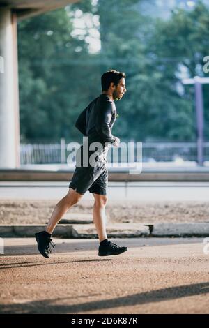 Athletic Young man in Sports Outfit ist Joggen in der Straße. Er läuft in einer urbanen Umgebung unter einer Brücke mit Autos im Hintergrund. Stockfoto
