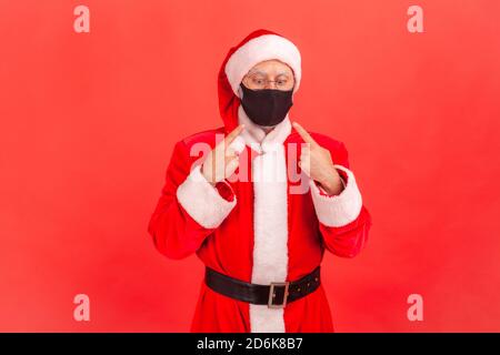 Erschrocken lustigen Mann in santa claus Kostüm zeigt Finger und versucht, auf schützende hygienische Maske auf seinem Gesicht, müde von covid-19 Epidemie zu suchen. Indoo Stockfoto