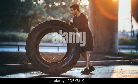 Starker muskulöser Fit junger Mann macht Übungen auf der Straße. Er hebt und rollte einen großen schweren Reifen in einer städtischen Umgebung. Stockfoto