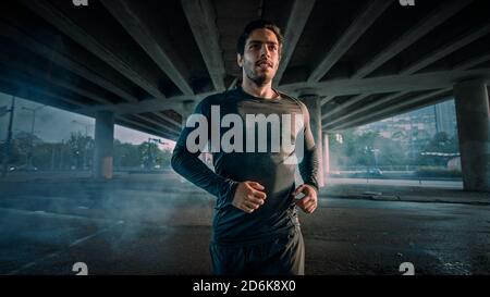 Portrait Aufnahme eines athletischen muskulösen jungen Mannes im Sport-Outfit Joggen auf der Straße. Er läuft in einer urbanen Umgebung unter einer Brücke. Stockfoto
