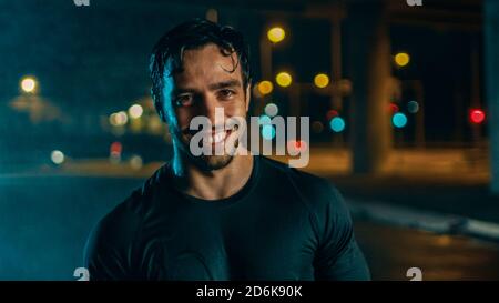 Portraitaufnahme eines sportlichen jungen Mannes im dunklen Sport-Outfit, der nach einem Street Workout an einem regnerischen Tag ein Lächeln auf den Kopf hat. Er ist in einem Abend Urban Stockfoto