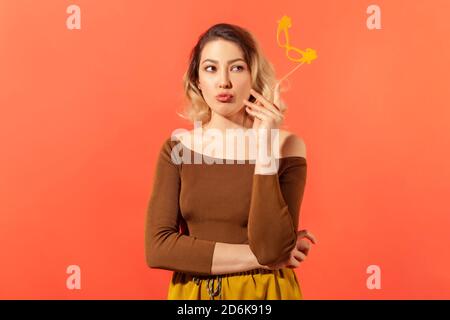 Attraktive nachdenkliche Frau hält Papiermaske auf Stick Nachdenken über ihr Kostüm auf Phantasie Party, träumen. Innenaufnahme des Studios isoliert auf Orang Stockfoto