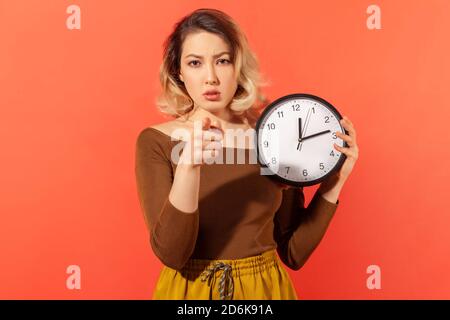 Zeit zum Handeln. Seriöse Frau hält große Wanduhr zeigt Finger auf Sie, Motivation, Warnung über Frist. Innenaufnahme des Studios isoliert auf orangefarbenem BA Stockfoto