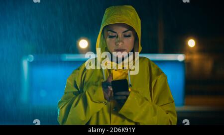 Schöne konzentrierte junge Mädchen in einem gelben Regenmantel ist mit einem Smartphone im Regen. Das Wetter ist regnerisch und es ist Nacht. Stockfoto