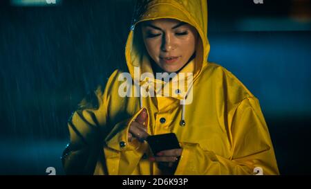 Schöne konzentrierte junge Mädchen in einem gelben Regenmantel ist mit einem Smartphone im Regen. Das Wetter ist regnerisch und es ist Nacht. Stockfoto