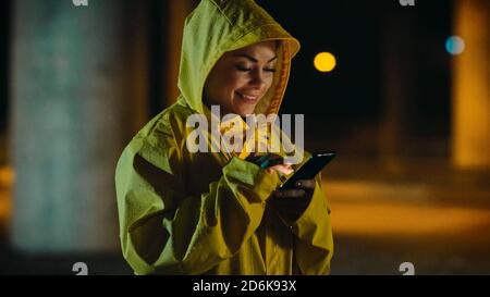 Schöne junge Mädchen in einem gelben Regenmantel ist mit einem Smartphone im Regen. Sie lächelt, nachdem sie eine Benachrichtigung erhalten hat. Wetter ist regnerisch und es ist Stockfoto