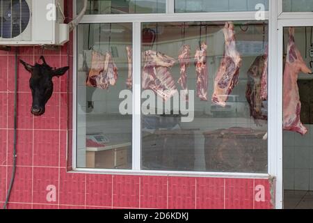 Blick durch schmutziges verschwommenes Fensterglas auf frische Fleischkadaver, die in der Straßenschlachterei auf dem kaukasus-Dorfmarkt hängen und den Kopf abtrennen. Stockfoto