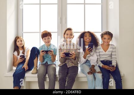 Glückliche multiethnische Kinder spielen Spiele auf Handys sitzen auf Fensterbank Stockfoto