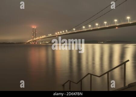 Humber Brücke hell mit Lichtern und Leuchtfeuer mit bewölktem Himmel und fließenden Mündung unten bei Flut bei Tagesanbruch in Hessle, Yorkshire, UK. Stockfoto