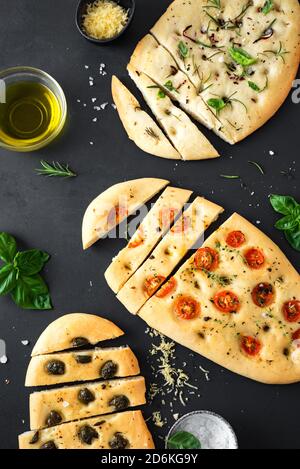 Italienisches Focaccia-Brot mit verschiedenen Füllungen auf schwarzem Hintergrund. Frisch gebackene Focaccia mit Tomaten, Oliven, Knoblauch und Kräutern, Draufsicht. Stockfoto