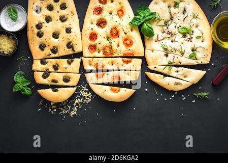 Italienisches Focaccia-Brot mit verschiedenen Füllungen auf schwarzem Hintergrund. Frisch gebackene Focaccia mit Tomaten, Oliven, Knoblauch und Kräutern, Draufsicht, Kopierraum. Stockfoto