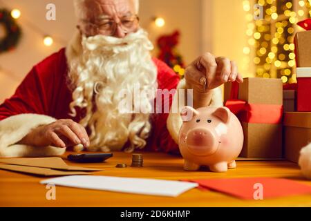 Der moderne Weihnachtsmann versucht, etwas Geld zu sparen und Putting Münzen im Sparschwein sitzen am Schreibtisch zu Hause Stockfoto