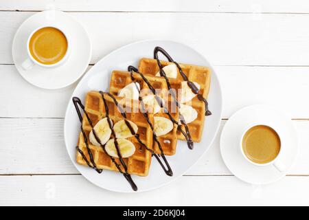 Hausgemachte Waffeln mit Bananenscheiben mit Schokolade und zwei Tassen Kaffee auf weißem Holztisch. Draufsicht. Stockfoto