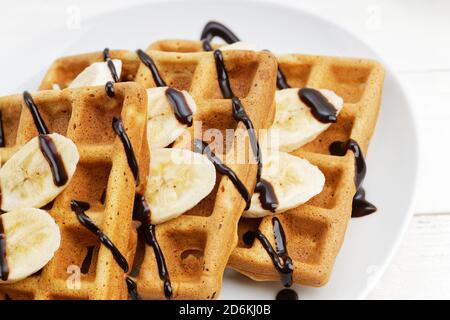 Nahaufnahme hausgemachte belgische Waffeln mit Bananenscheiben gekrönt mit Schokolade auf weißem Holztisch. Geringer Fokus. Stockfoto