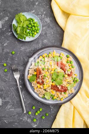 Quinoa-Haferbrei mit grüner Erbse, Mais und getrockneten Tomaten auf Keramikplatte auf grauem Betongrund und gelbem Textil. Draufsicht, flach liegend, nahe U Stockfoto