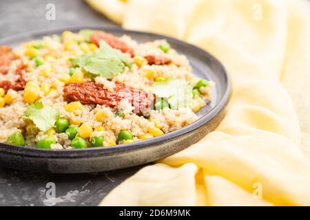 Quinoa-Haferbrei mit grüner Erbse, Mais und getrockneten Tomaten auf Keramikplatte auf grauem Betongrund und gelbem Textil. Seitenansicht, Nahaufnahme, auswählen Stockfoto