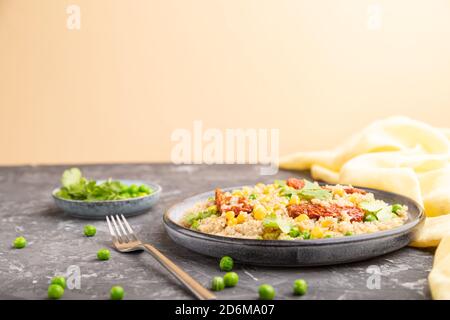 Quinoa-Haferbrei mit grüner Erbse, Mais und getrockneten Tomaten auf Keramikplatte auf grauem und orangefarbenem Hintergrund und gelbem Textil. Seitenansicht, Kopierbereich. Stockfoto