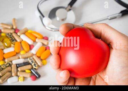 Hand drückt ein Gummiherz, ein Stethoskop auf einem Arzttisch und verstreute Pillen. Konzept von Gesundheit und Drogenmissbrauch Stockfoto