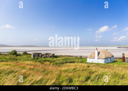 Traditionell gebaute weiße Hütte mit Strohdach, neben der türkisfarbenen Bucht, mit stürmischen bewölkten dunklen Himmel über.Insel North Uist, Schottland Stockfoto