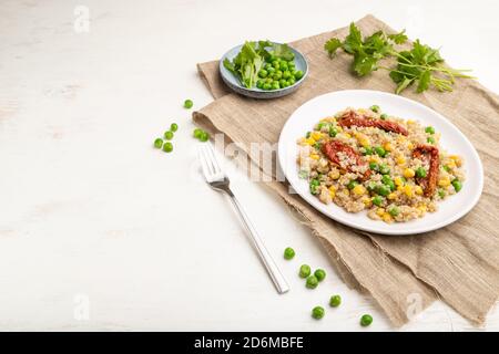 Quinoa-Haferbrei mit grüner Erbse, Mais und getrockneten Tomaten auf Keramikplatte auf weißem Holzhintergrund und Leinentextilien. Seitenansicht, Kopierbereich. Stockfoto