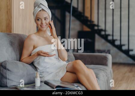 Fröhliche Frau von europäischem Aussehen gilt Gesichtscreme für gesunde weiche Haut, sitzt auf einem bequemen Sofa, umgeben von einer Flasche Lotion und Magazin, Stockfoto