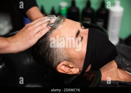 Weißer kaukasischer Mann mit schwarzer Maske des Schutzes Pandemie Coronavirus Legt sich, während die Hände eines Barbiers seine Haare waschen Mit Wasser und Seife in ha Stockfoto