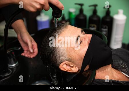 Weißer kaukasischer Mann mit schwarzer Maske des Schutzes Pandemie Coronavirus Legt sich, während die Hände eines Barbiers seine Haare waschen Mit Wasser und Seife in ha Stockfoto