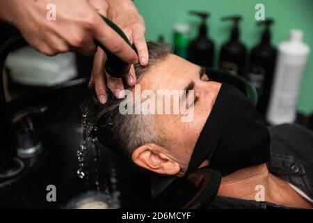 Weißer kaukasischer Mann mit schwarzer Maske des Schutzes Pandemie Coronavirus Legt sich, während die Hände eines Barbiers seine Haare waschen Mit Wasser und Seife in ha Stockfoto