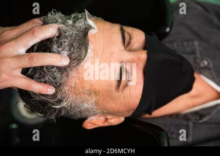 Weißer kaukasischer Mann mit schwarzer Maske des Schutzes Pandemie Coronavirus Legt sich, während die Hände eines Barbiers seine Haare waschen Mit Wasser und Seife in ha Stockfoto