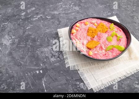 Hummus mit Rüben und mikrogrünen Basilikum Sprossen in Keramikschale auf schwarzem Beton Hintergrund und Leinentextilien. Seitenansicht, Kopierbereich. Stockfoto