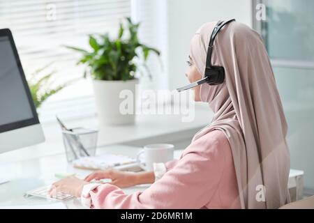 Moderne junge muslimische weibliche Bedienerin in Hijab und Headset-Typisierung Im Büro Stockfoto