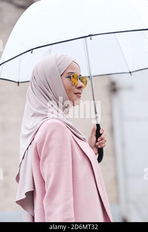Junge stilvolle muslimische Frau in Hijab, Sonnenbrille und Strickjacke hält Regenschirm Stockfoto
