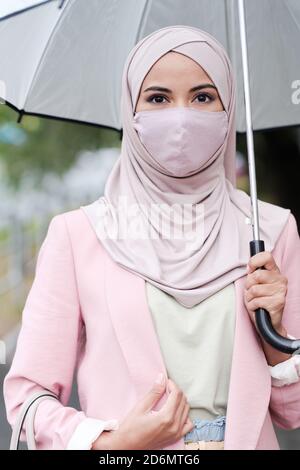 Junge muslimische Frau in Schutzmaske unter Regenschirm auf stehen Regentag Stockfoto