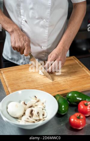 Hände des jungen männlichen Küchenchefs, der frische Zwiebeln und Champignons schneidet Auf Holzbrett Stockfoto