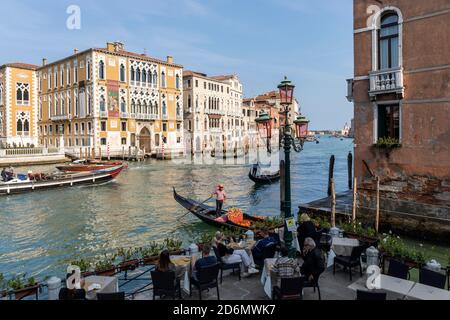 Touristen genießen die Aussicht von einem Restaurant am Kanal auf Palazzo Cavalli-Franchetti und Palazzo Barbaro. Gondelbahn (Gondoliere) auf dem Canal Grande, Venedig, Stockfoto