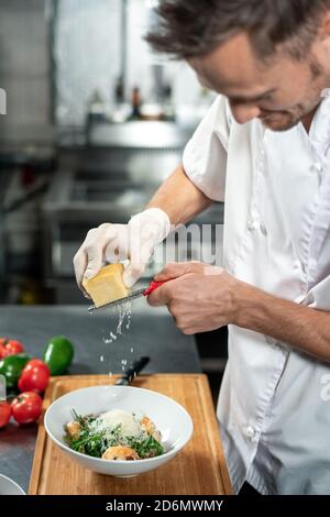 Professioneller junger Koch in einheitlichem Käsereibenraspeln in Keramikschale Mit Salat Stockfoto