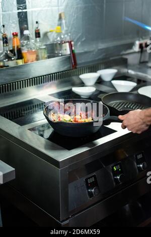 Hand des jungen Küchenchefs Gießen halten Bratpfanne mit gehackt Gemüse Stockfoto