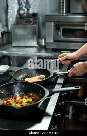Hände des männlichen Küchenchefs rösten ein Stück Lachs beim Kochen Fisch und Gemüse Stockfoto
