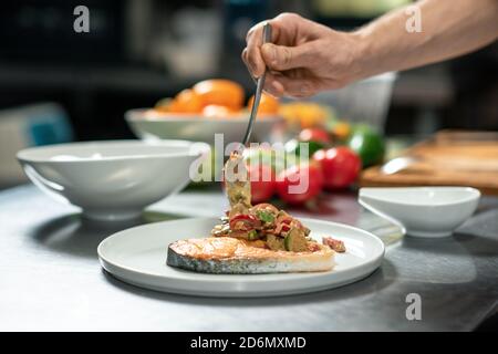 Hand des jungen männlichen Küchenchefs, der gebratenes Gemüse auf Stück setzt Von gebratenem Lachs Stockfoto