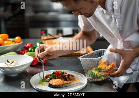 Junger männlicher Koch verzierend gebratenes Gemüse auf Stück gebraten Lachs auf dem Teller Stockfoto