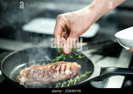 Hand des Küchenchefs setzen grüne aromatische Kräuter auf gerösteten Stück Von Rindfleisch in der Pfanne Stockfoto