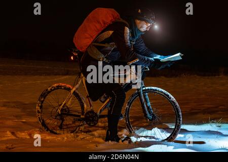 Nachtfahrerin überprüfen den Streckenverlauf im Licht eines Scheinwerfers. Verlorener Weg in Schneeverwehung, Schneeflocken schmelzen auf dunklen Offroad-Reifen. Extreme-Bike Stockfoto