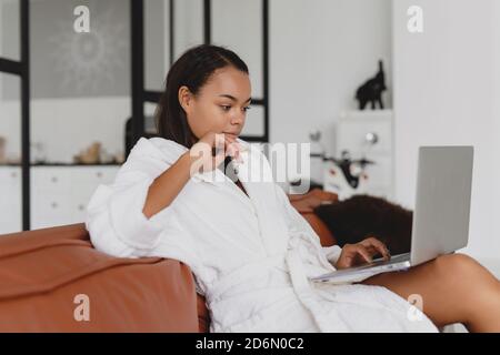 Attraktive afroamerikanische Frau im weißen Bademantel mit Laptop, während sie auf einer Couch zu Hause sitzt. Stockfoto