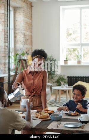 Junge glückliche Frau in Casualwear Gießen Kaffee oder Tee in Tasse ihres Mannes Stockfoto