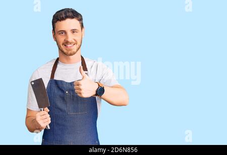 Schöner junger Mann mit Bär trägt professionelle Schürze halten Messer lächelnd glücklich und positiv, Daumen nach oben tun ausgezeichnet und Zustimmung Zeichen Stockfoto