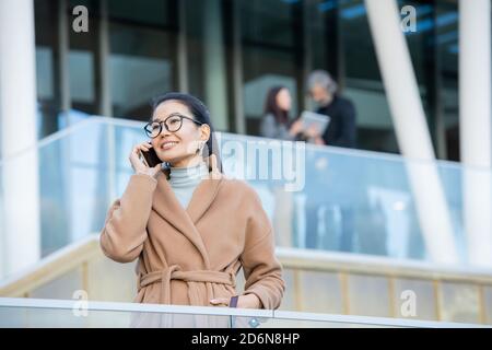 Glückliche junge asiatische Geschäftsfrau im beigen Mantel im Gespräch auf mobile Telefon im Freien Stockfoto