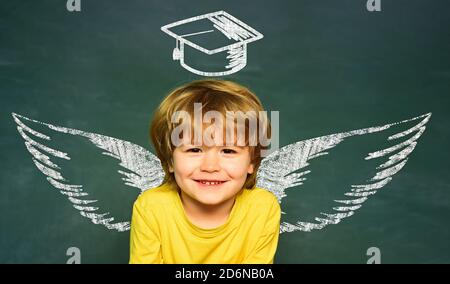 Schulflügel und Traum. Frohe Schüler. Schulkonzept. Schulungsraum. Lustiger kleiner Junge, der auf Tafel zeigt. Schulkinder gegen grüne Tafel Stockfoto