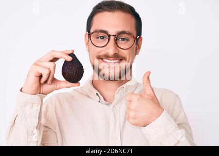 Junge gutaussehende Mann hält Avocado lächelnd glücklich und positiv, Daumen nach oben tun ausgezeichnet und Zustimmung Zeichen Stockfoto